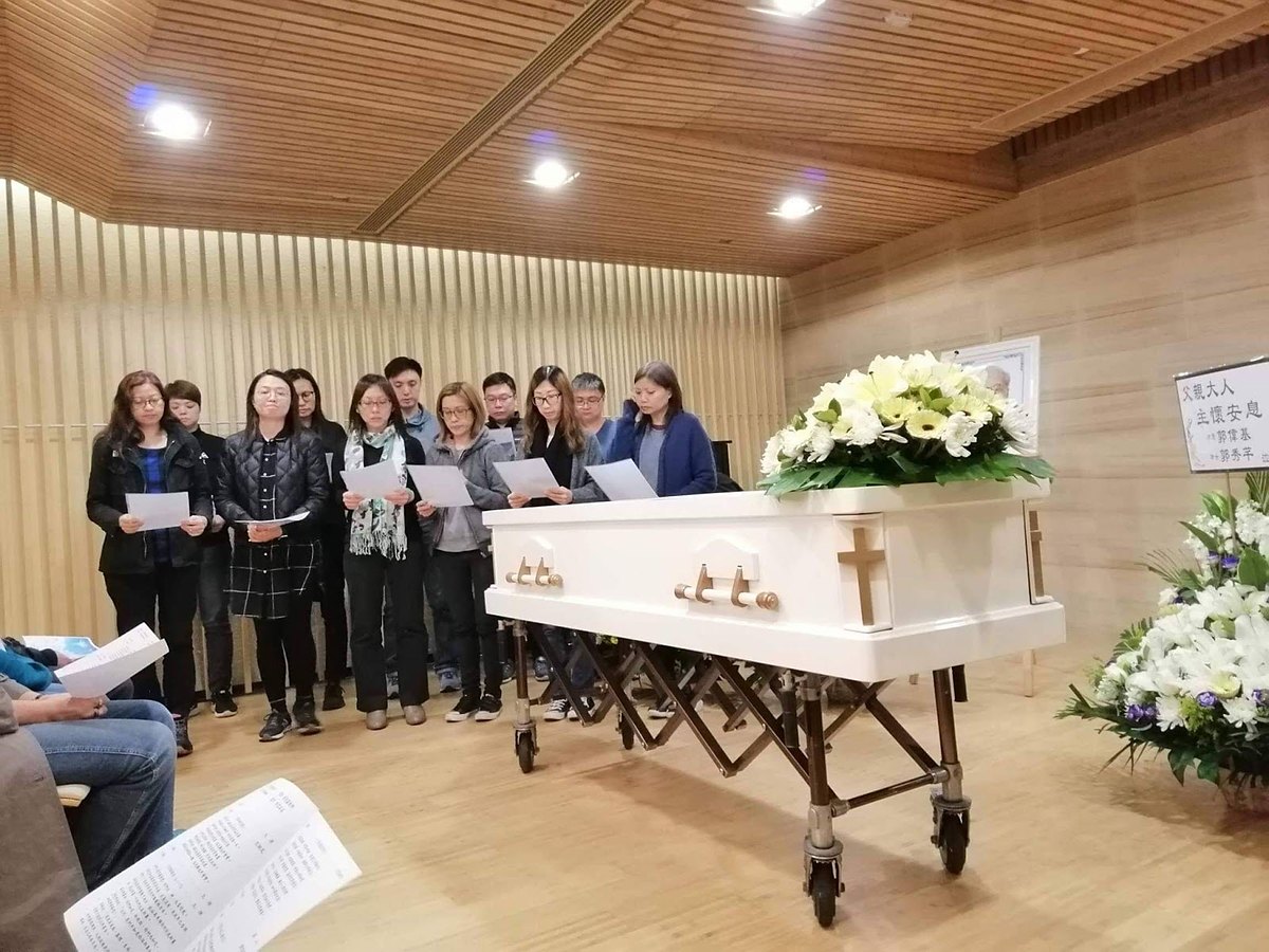 The memorial service: family standing together holding hymn sheets beside a white coffin with a cross and white flowers.